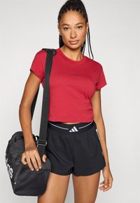 Κόκκινο κομμένο t-shirt με κοντά μανίκια και διακριτικό λογότυπο, συνδυασμένο με μαύρο σορτς που διαθέτει λαστιχένια ζώνη με λογότυπο και μαύρο αθλητικό τσαντάκι.