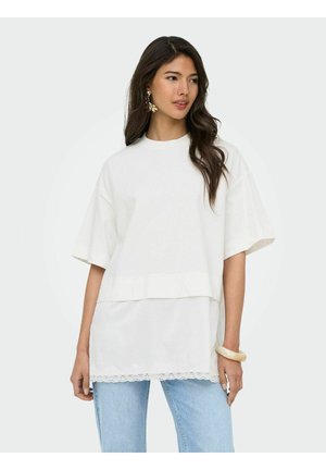 Femme aux longs cheveux foncés portant un haut blanc superposé à manches courtes avec un ourlet en dentelle et un jean bleu clair, accessoirisée avec des boucles d’oreilles dorées et un bracelet.