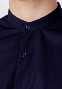 Camicia blu navy con colletto alla cinese, dotata di chiusura con un solo bottone e una texture di tessuto liscia. Design semplice e moderno senza fantasie.