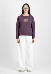 Lilla sweatshirt med et logo, rund hals, og ribbede manchetter, parret med hvide brede bukser og grå sneakers med en gennemsigtig sål.
