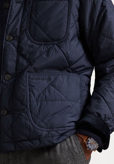 Jūras spēku krāsas quiltēta jakas ar pogu aizdari, ar lielu kabatu un ribotām piedurknēm. Metāliski akcenti un gluda tekstūra.