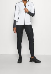 Witte zip-up jack met grijze accenten en logo op de mouwen, gecombineerd met zwarte strakke leggings met een glanzend patroon. Zwarte sportschoenen.