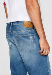 Svijetloplavi traperice s oštećenjima, kožna oznaka s natpisom "Calvin Klein Jeans" i srednji struk.