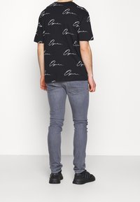 Camiseta negra con patrón de firmas blancas, corte holgado; combinada con jeans grises ajustados y zapatillas negras; fondo liso.