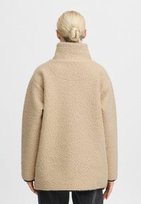 Beige fleecejakke med høj krave, løs pasform og en tekstureret, blød overflade. Har lange ærmer og et enkelt, minimalistisk design.