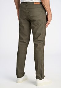 Pantalon ajusté vert olive en tissu texturé, à cinq poches, avec une étiquette en patch au dos. Porté avec des baskets blanches.