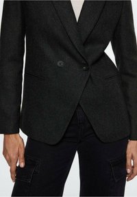 Zwarte visgraat blazer met een enkele knoopsluiting, geknipte revers en zijzakken, gecombineerd met donkere cargobroeken.