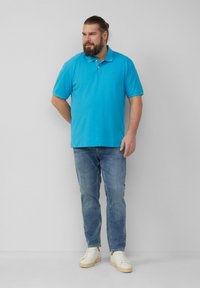 Polo turquoise avec un col, associé à un jean bleu clair et des baskets blanches. La chemise présente un logo discret sur la poitrine.