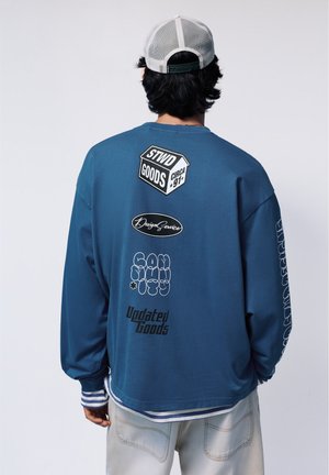 Personne portant un t-shirt à manches longues bleu avec du texte et des graphiques imprimés en blanc et noir dans le dos, ainsi qu'une casquette de baseball grise en mesh.