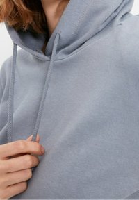Sudadera gris claro con cordones, de textura de tela suave y un diseño relajado. Vista en primer plano de la capucha y una mano sosteniéndola.
