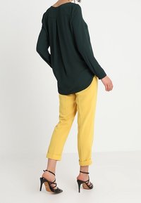 Femme portant un chemisier à manches longues vert foncé, un pantalon jaune retroussé et des sandales à talons hauts noires à lanières, debout devant un fond uni.
