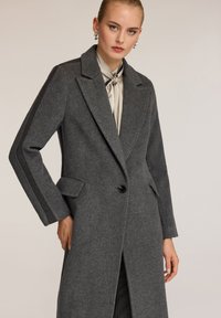 Cappotto grigio di lana con chiusura a un solo bottone, colletto a revers, tasche laterali e strisce lungo le maniche; texture liscia.