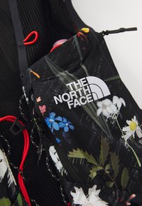 Svart ryggsäck från North Face med blommönster, synlig vit logotyp och röda dragsnören på en svart nätbakgrund.