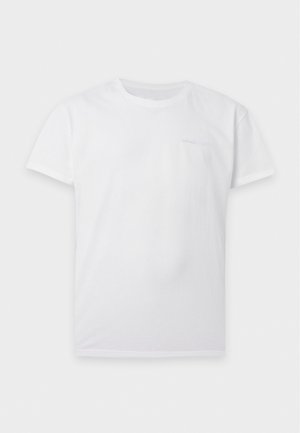 Un homme pose avec un tee-shirt blanc, un short en jean bleu, des chaussettes blanches et des baskets blanches sur un fond clair et uni.