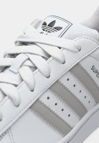 Close-up of white Adidas Superstar sneaker with gray stripes, white laces, and Adidas trefoil logo on the tongue.