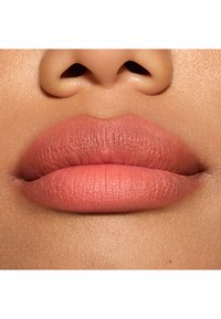 Gros plan sur des lèvres pulpeuses avec une finition mate corail-rosé, montrant des détails de texture fine et un arrière-plan de teint naturel.