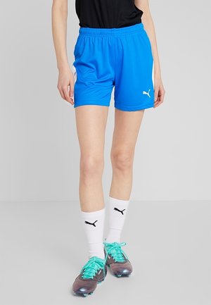 Persoon draagt felblauwe Puma sportshort, witte Puma sokken en donkere voetbalschoenen met turquoise veters, staand op een grijze vloer.