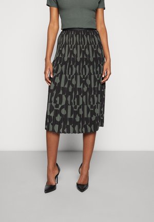 Femme portant une jupe midi plissée noire et vert olive avec des motifs abstraits, associée à des chaussures à talons hauts noires et un top vert foncé.
