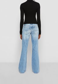 Henkilö, jolla on päällään vaaleansiniset leveät farkut ja tiukka musta pitkähihainen paita, seisoo yksinkertaista vaaleanharmaata taustaa vasten.