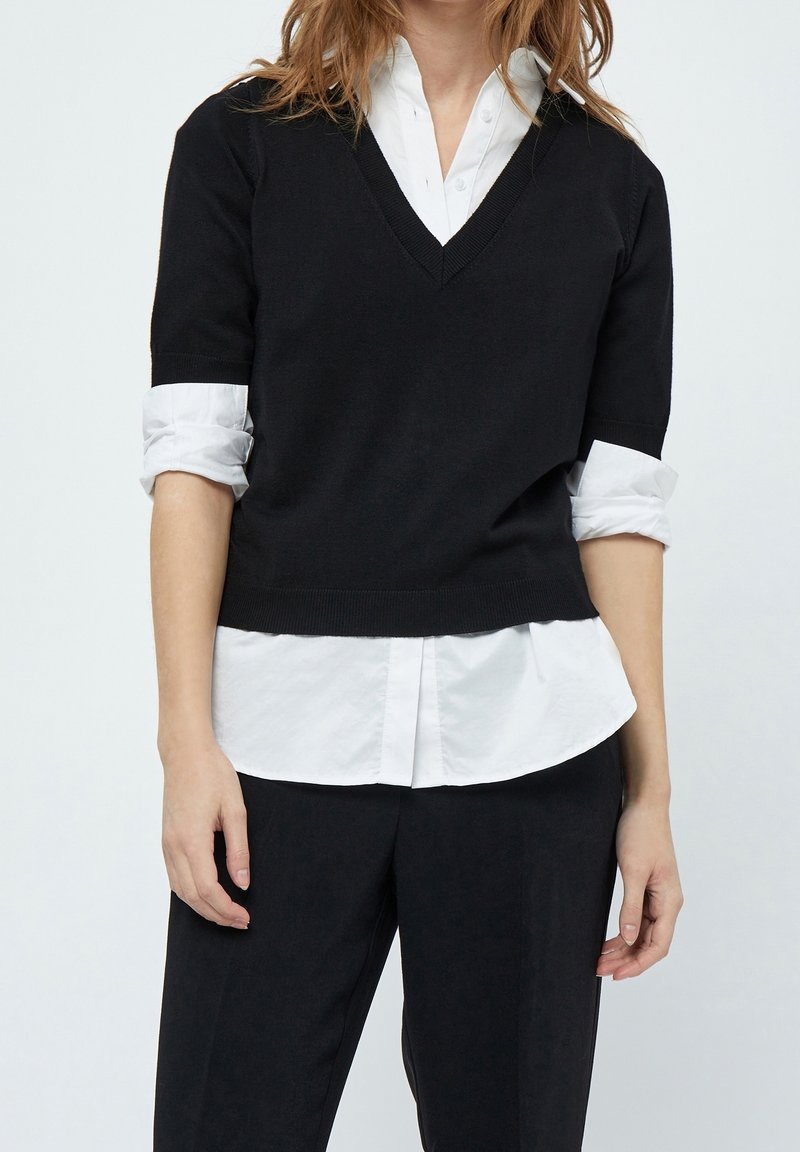 Femme portant un pull noir à col en V et manches courtes sur une chemise blanche boutonnée aux manches retroussées, avec un pantalon noir, debout devant un fond neutre.