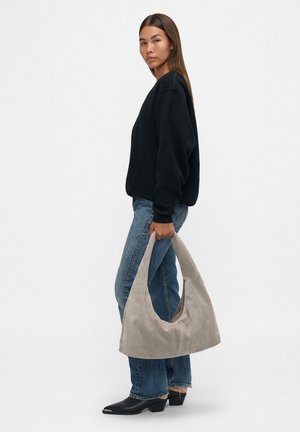 Borsa hobo in suede grigio chiaro, con una forma curvata e un'ampia apertura. Il modello indossa una felpa nera e jeans blu per contrasto.