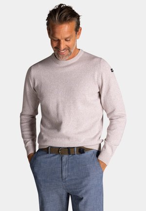 Man met kort haar en baard, gekleed in een lichtbeige trui met ronde hals en blauwe broek met een gevlochten bruine riem, handen in de zakken.