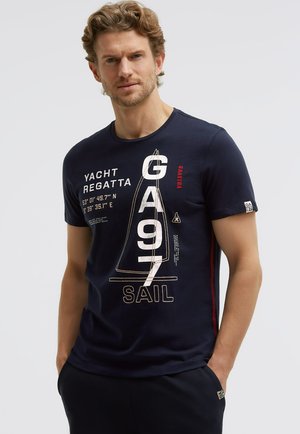 Mann mit kurzen lockigen Haaren, der ein marineblaues T-Shirt mit weißem und rotem Yachtregatta- und Segelmotiv trägt, steht mit den Händen in den Taschen.