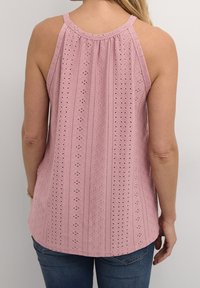 Donna che indossa un top halter senza maniche rosa con pizzi e motivi geometrici, abbinato a jeans blu, in piedi davanti a uno sfondo bianco semplice.