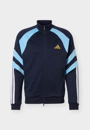 Ναυτικό τζάκετ με ανοιχτόχρωμες μπλε πλάγιες πλάκες στους ώμους, δύο διαγώνιες ρίγες στα μανίκια, λευκές ρίγες κατά μήκος των χεριών και μικρό κίτρινο λογότυπο στο στήθος.