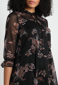 Robe noire à fleurs en tissu transparent, avec un col rond à nouer, des manches trois-quarts et une coupe décontractée avec des motifs floraux variés.