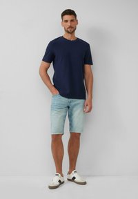 Marineblauwe T-shirt met korte mouwen gecombineerd met lichtblauwe denim shorts en witte sneakers met donkere accenten, staande tegen een effen achtergrond.