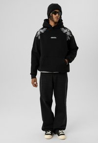 Zwart hoodie met bloemenborduurwerk op de schouders, voorzak met kangoeroe-ontwerp, gecombineerd met ruime zwarte broek en zwarte sneakers.