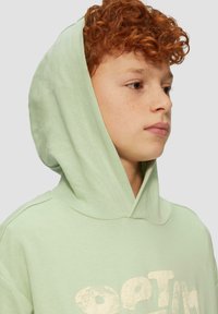Haut en coton doux de couleur vert clair, doté d'une poche kangourou et d'un grand graphique texturé avec l'inscription "OPTIM". Comprend une capuche avec cordon de serrage.