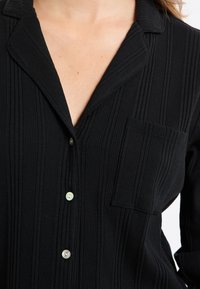 Czarna prążkowana koszula zapinana na guziki z kołnierzykiem, pojedynczą kieszonką i dwoma jasnymi guzikami, prezentująca teksturowany materiał.