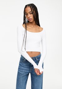 Bílý žebrovaný dlouhý rukáv crop top s otevřeným výstřihem, párovaný s modrými širokými džínami s vysokým pasem. Materiál vypadá jemně a lehce.