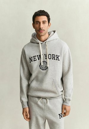 Mann mit kurzen lockigen Haaren trägt einen hellgrauen Hoodie und Jogginghose mit New York Schriftzug und steht vor einem schlichten Hintergrund.