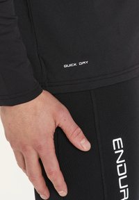 Svart snabbtorkande träningströja med synlig sömnad, hand vilande nära sömmen. Vita textaccenter "ENDURE" och "QUICK DRY" på tyget.