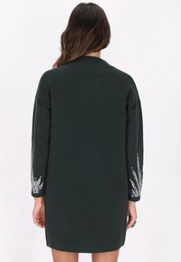 Dunkelgrünes Strickkleid mit langen Ärmeln, das über filigrane silberne Stickereien an den Ärmeln verfügt. Gerader Schnitt mit entspannter Silhouette.