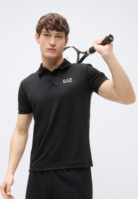Svart polotrøye med strukturert krage, korte ermer og EA7-logo på venstre bryst. Modell holder en tennisracket. Enkelt design.