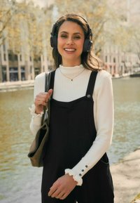 Zwarte overall jurk over een witte geribbelde coltrui; zwarte hoofdtelefoon, gerimpelde mouwen en een bruine schoudertas; buitensetting met water op de achtergrond.