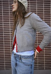 Junge Frau trägt eine graue Strickjacke über roten und blauen Oberteilen, blaue Jeans und eine weiße Strickmütze, steht vor einer gefliesten Wand.