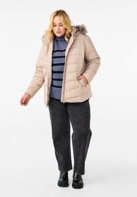 Beige podšívková bunda s kapucí na kožíšku, se zapínáním na zip a volným střihem. Nosená přes modro-černý pruhovaný svetr, v kombinaci s tmavými džíny.