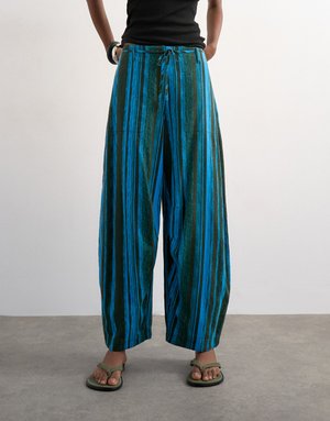 Personne portant un pantalon ample à jambes larges avec des rayures verticales bleues et vertes, assorti à des sandales vertes et un haut sans manches noir.