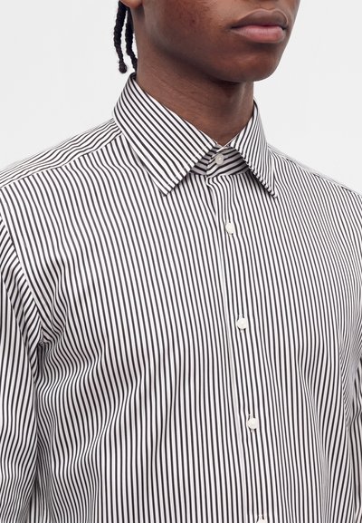 Jeune personne portant une chemise boutonnée à rayures verticales noires et blanches avec col, face à un fond clair uni.