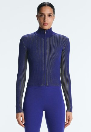 Woman wearing a fitted, ribbed, long-sleeve zip-up sweater and matching high-waist pants in deep blue with subtle vertical stripes.