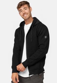 Black zip-up hoodie with a drawstring hood, ribbed cuffs, a front pocket, and a small patch detail on the left sleeve. Worn over a white t-shirt.