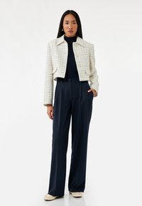 Crème tweed cropped jasje met kleine zakken en knoopsluitingen, gecombineerd met marineblauwe high-waisted broeken. Lichtgewicht stof, gestructureerd ontwerp.