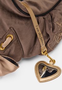 Bolsa de mão em camurça castanha com detalhes em dourado, apresentando um charme em forma de coração com espelho e uma alça bege inscrita com "Eu sou a mais bela."