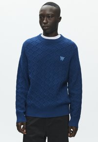 Pull en tricot bleu avec un motif en losanges, poignets et ourlet côtelés, et un petit logo sur la poitrine. Porté par-dessus un t-shirt blanc à col rond.