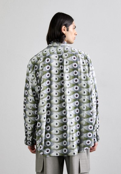 Chemise à manches longues, coupe ample, arborant un motif d'yeux répété en vert, gris et blanc, confectionnée dans un tissu lisse avec des détails de boutons.